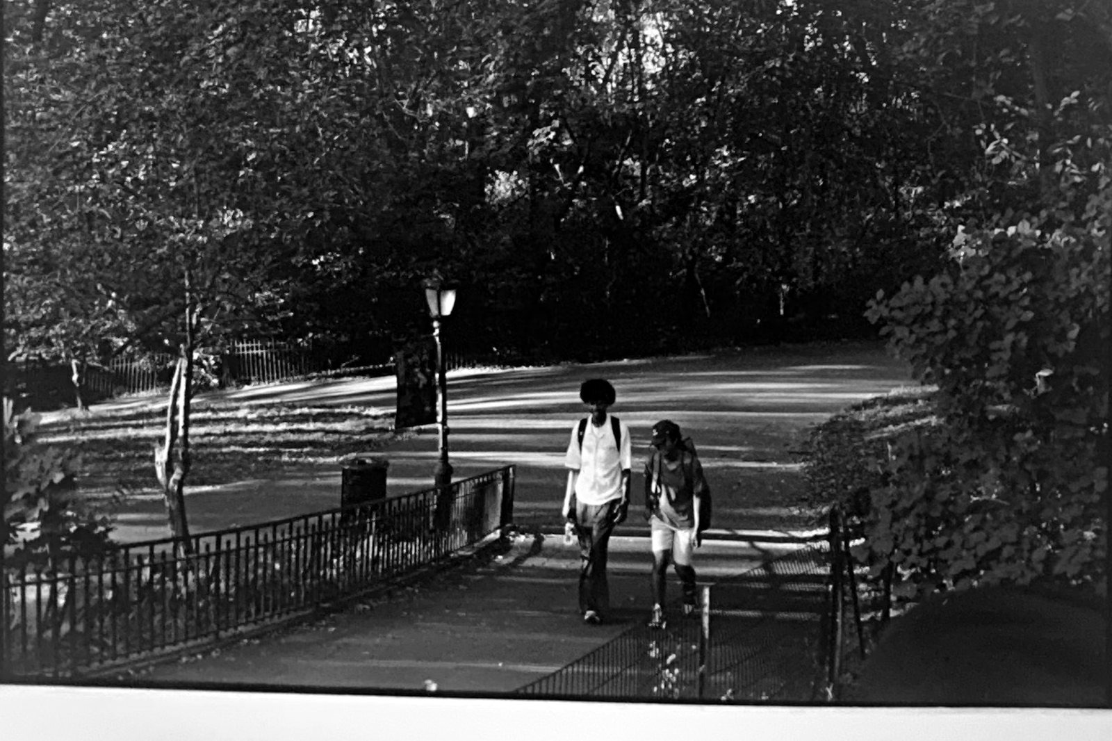 Black and white photograph of two people walking a paved path past a park fence.