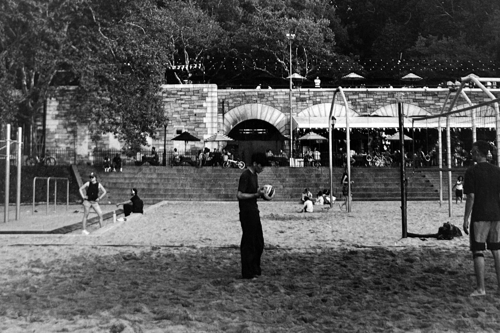 Black and white photograph of a sand volleyball court with players and market awnings behind.