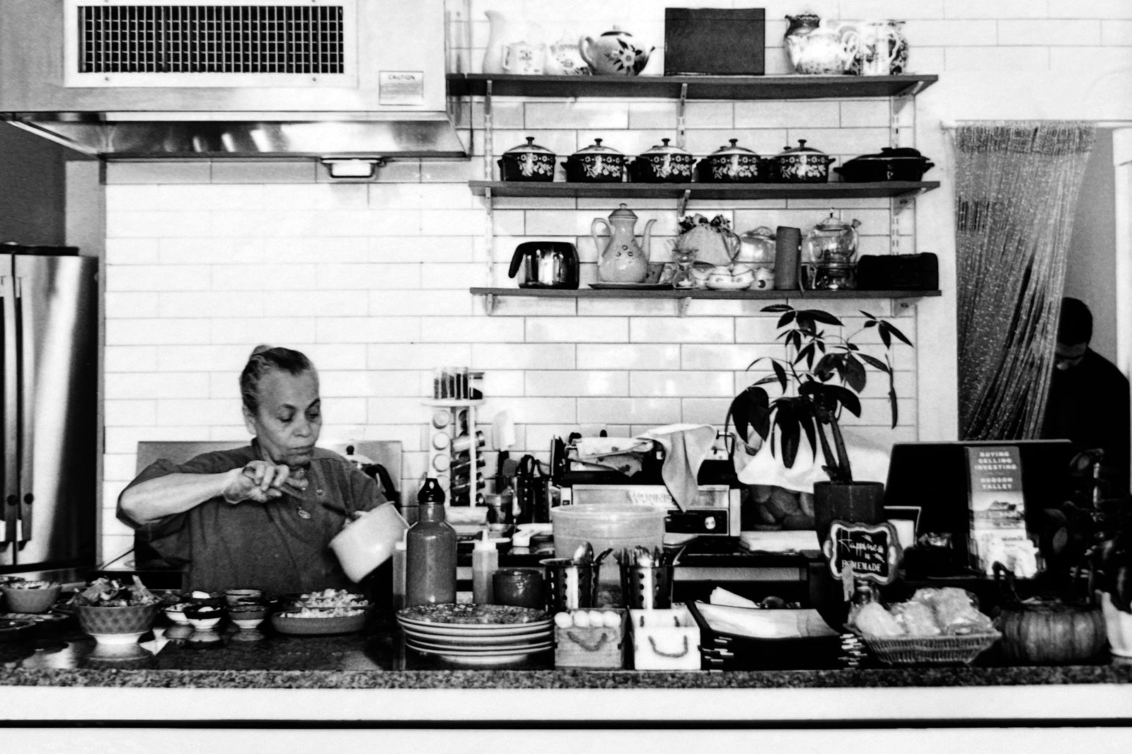 Black and white photograph of a cook working behind the counter of a tiled kitchen.